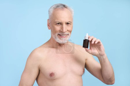 Téléchargez les photos : Beau Blanc cheveux gris barbu homme âgé montrant un bocal avec sérum anti-âge, souriant en regardant la caméra, isolé sur fond bleu. Beauté masculine. Rajeunissant anti rides visage soin de la peau - en image libre de droit