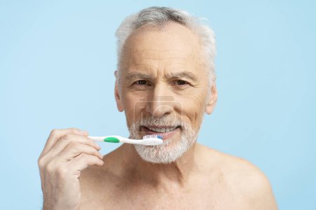 Photo for Attractive senior man looking in mirror, brushing teeth with toothbrush isolated on blue background. Concept of dental care and health - Royalty Free Image