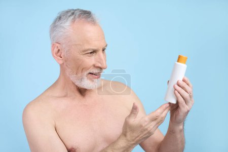 Téléchargez les photos : Homme âgé barbu aux cheveux gris caucasiens, tenant une bouteille blanche avec de la lotion solaire, souriant tout en posant avec un torse nu, isolé sur fond bleu. Soins de la peau, protection solaire et concept d'été - en image libre de droit