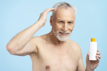 Téléchargez les photos : Portrait d'un homme âgé barbu et souriant tenant une bouteille de shampooing, touchant les cheveux, regardant la caméra. Homme retraité attrayant avec torse nu, posant pour l'image. Soins capillaires, concept de perte de cheveux - en image libre de droit
