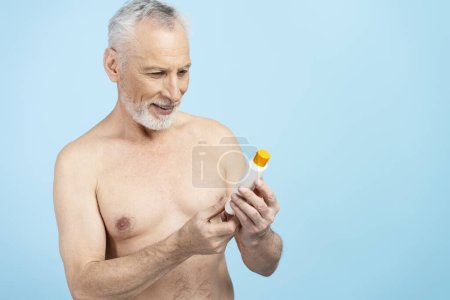 Photo for Portrait of erderly shirtless man holding sunscreen cream after shower isolated on blue background. Healthy lifestyle, skin care - Royalty Free Image