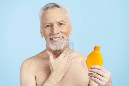 Téléchargez les photos : Attrayant souriant senior homme appliquant crème solaire après la douche isolé sur fond bleu. ? oncept des soins du corps - en image libre de droit