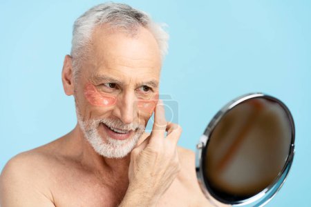 Photo for Good looking senior gray haired man with patches under eyes with naked torso, holding mirror after shower, looking in mirror. Pensioner doing cosmetology procedures. Morning routine, skin care - Royalty Free Image