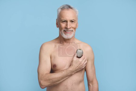 Photo for Happy senior man holding a pacemaker over his naked torso, smiling, confidently looking at camera, standing shirtless, isolated on blue background. Healthcare and medicine. Cardiac surgery concept - Royalty Free Image