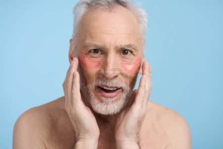 Photo for Close-up Caucasian gray haired bearded senior man 60 years old, with anti puffiness and anti dark circles patches under eyes, holding his hands on face, looking at camera, isolated on blue background - Royalty Free Image