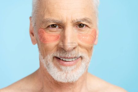 Téléchargez les photos : Portrait d'un beau retraité âgé avec des taches sous les yeux isolés sur fond bleu. Concept de soin de la peau - en image libre de droit