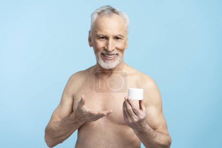 Foto de Retrato de un hombre mayor de pelo gris barbudo sonriente con torso desnudo, sosteniendo crema, aislado sobre fondo azul. Concepto de cuidado de la piel - Imagen libre de derechos