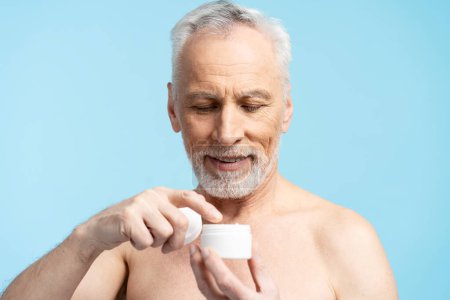 Téléchargez les photos : Portrait d'un homme âgé souriant au torse nu, appliquant de la crème, isolé sur fond bleu. Concept de soins de la peau - en image libre de droit