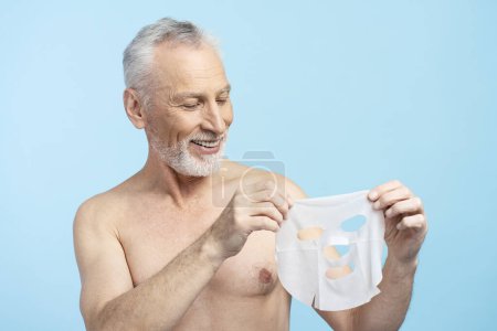 Téléchargez les photos : Portrait de séduisant homme âgé avec torse nu, tenant masque face à face, isolé sur fond bleu. Mode de vie sain, soin de la peau - en image libre de droit