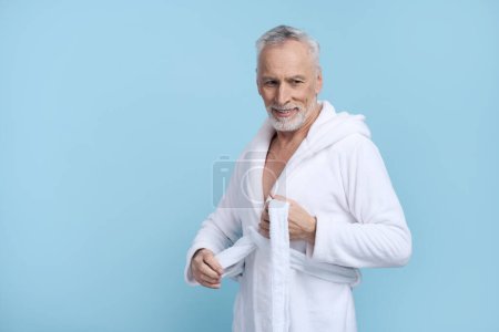 Téléchargez les photos : Homme âgé barbu aux cheveux gris caucasiens confiants, sourit en regardant de côté, met un peignoir éponge blanc, exprimant des émotions positives, isolé sur fond bleu avec un espace publicitaire de copie. Masculin spa beauté - en image libre de droit