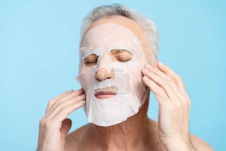 Téléchargez les photos : Portrait de séduisant homme âgé avec torse nu, appliquer un masque sur le visage, isolé sur fond bleu. Mode de vie sain, soin de la peau - en image libre de droit