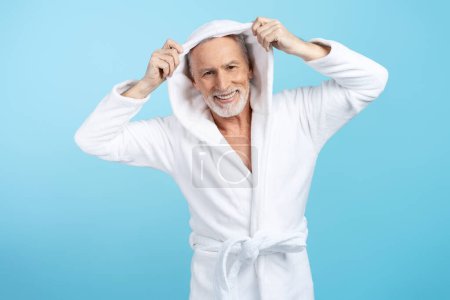 Téléchargez les photos : Beau mâle âgé en peignoir vêtu d'une capuche, regardant la caméra isolée sur fond bleu. Concept de routine matinale, procédures beauté - en image libre de droit