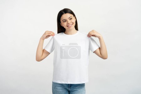 Foto de Retrato de una linda adolescente sonriente con una camiseta blanca apuntándose a sí mismo mirando hacia otro lado aislado sobre un fondo blanco. Burla. - Imagen libre de derechos