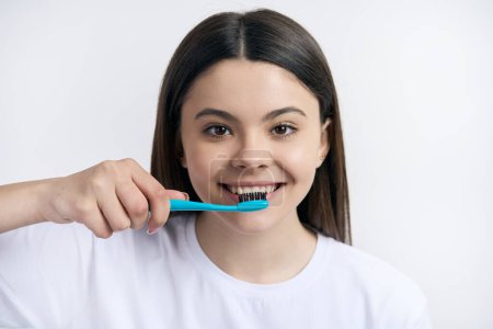 Foto de Headshot encantadora chica adolescente caucásica consciente en camiseta blanca casual, cepillarse los dientes con cepillo de dientes, aislado en el fondo del estudio blanco. Salud y medicina. Odontología pediátrica. Cuidado oral - Imagen libre de derechos