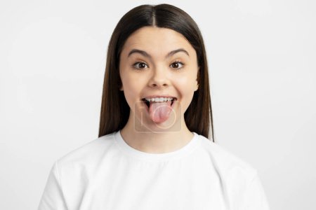 Téléchargez les photos : Portrait d'adolescente drôle et joyeuse montrant la langue regardant la caméra isolée sur fond blanc. Concept de style de vie positif - en image libre de droit