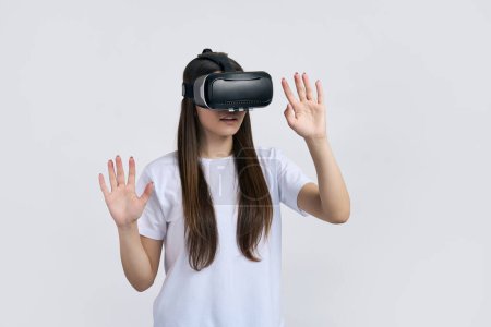 Téléchargez les photos : Teenager girl en t-shirt blanc maquette, l'expérience du jeu de réalité virtuelle avec des lunettes VR, geste avec les mains, fond blanc isolé. Personnel Divertissement Technologie Innovation Simulation Hi-Tech - en image libre de droit