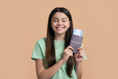 Photo for Caucasian happy pretty teenager traveler girl smiling cutely, holding boarding pass, waiting upcoming flight, enjoying happy summer holidays, posing with her eyes closed on beige isolated background. - Royalty Free Image