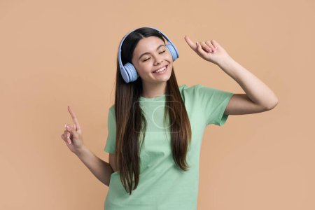 Photo for Caucasian cheerful happy smiling teen girl dancing, listening to music on blue wireless headphones. Lovely adolescent child have fun, points fingers while dancing over isolated beige color background - Royalty Free Image
