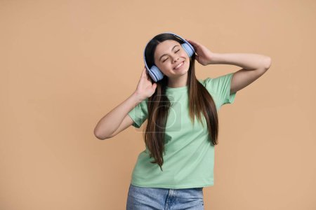 Photo for Charming Caucasian teenage girl in casual clothes, putting on blue wireless headphones, relaxing with her eyes closed while listening to music or cool soundtrack, over isolated beige color background - Royalty Free Image