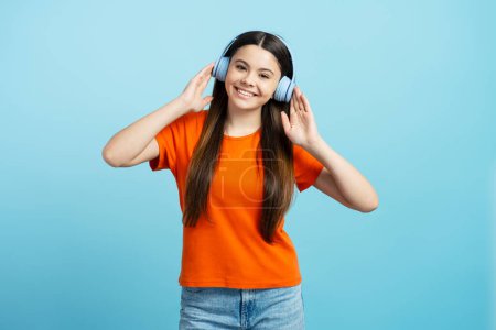 Photo for Happy smiling teenage girl wearing orange t shirt listening music in wireless headphones isolated on blue background. Concept of enjoying song - Royalty Free Image