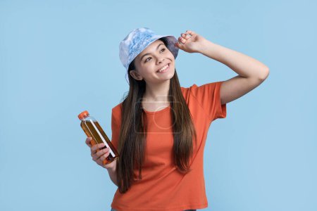 Foto de Feliz chica adolescente caucásica en camiseta naranja y elegante sombrero de panama azul, sonriendo con cuidado mirando hacia otro lado, sosteniendo una botella con refrescante bebida de limonada, aislado en el fondo de color azul. Concepto de verano - Imagen libre de derechos