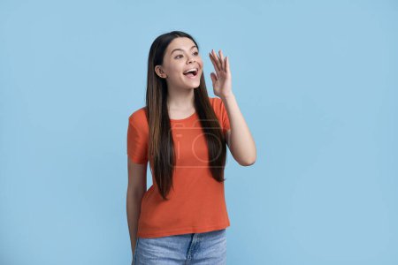 Photo for Smiling positive dark-haired stylish stunning beautiful Caucasian teenager, teen girl in orange t-shirt and blue jeans, holding her hand near her face, shouting over blue background. Copy space - Royalty Free Image