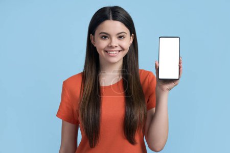 Foto de Encantadora chica adolescente caucásica sonriendo a la cámara, mostrando un teléfono móvil inteligente con pantalla táctil digital en blanco con espacio de copia para aplicaciones móviles o texto publicitario, aislado en color azul - Imagen libre de derechos