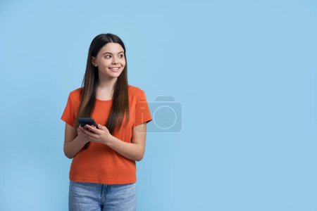 Foto de Preciosa chica adolescente caucásica en azul vaquero casual y camiseta naranja, charlando en línea en su teléfono inteligente moderno, escribiendo mensajes de texto, mirando soñadoramente a un lado, sonriendo sobre el fondo azul aislado - Imagen libre de derechos