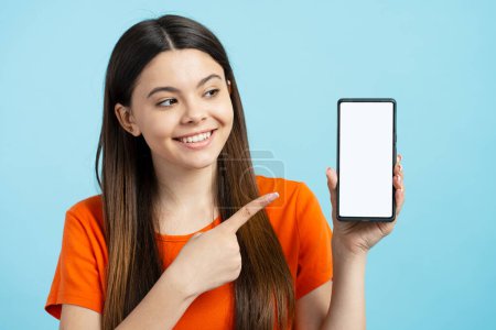 Foto de Sonriente adolescente con camiseta naranja sosteniendo el teléfono móvil apuntando con el dedo a la pantalla digital aislada sobre fondo azul, maqueta. Mujer feliz utilizando la aplicación móvil moderna de compras en línea - Imagen libre de derechos