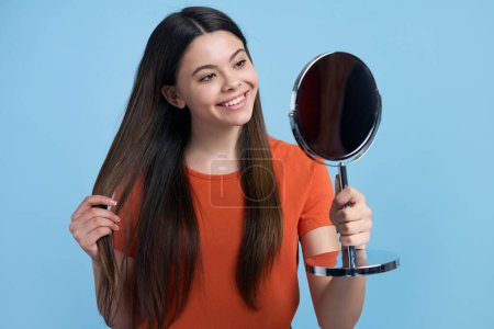Foto de Chica adolescente caucásica admirándose a sí misma, mirando al espejo cosmético, sonriendo, alisando su hermoso cabello largo, sintiendo felicidad y satisfacción por su apariencia, fondo de color azul aislado - Imagen libre de derechos