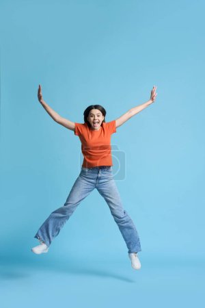 Téléchargez les photos : Portrait émotionnel conceptuel studio pleine grandeur d'une belle adolescente, portant un t-shirt orange et un jean bleu, souriant en regardant la caméra, sautant par-dessus un fond bleu isolé. Concept de personnes - en image libre de droit