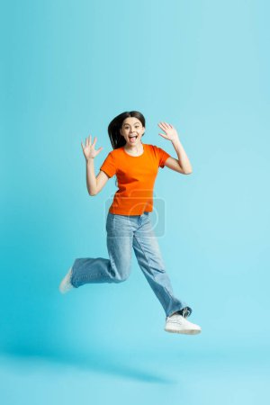Photo for Beautiful overjoyed teenage girl wearing stylish jeans, orange t shirt jumping high isolated on blue isolated background - Royalty Free Image