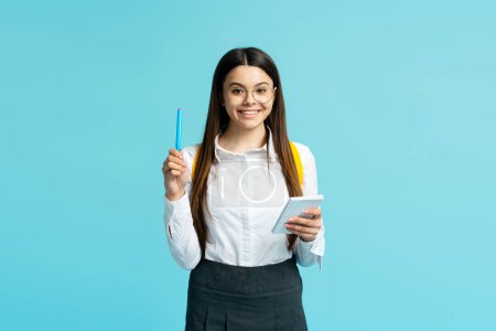 Photo for Portrait of cute smiling teenage girl wearing glasses and school uniform writing in notebook isolated on blue background. School concept - Royalty Free Image