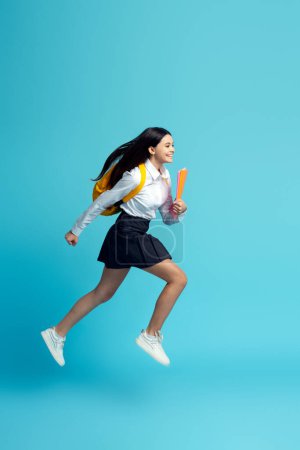 Foto de Chica adolescente feliz vistiendo uniforme, mochila, sosteniendo cuadernos corriendo rápido a la escuela aislado sobre fondo azul. Regreso a la escuela, concepto de educación - Imagen libre de derechos