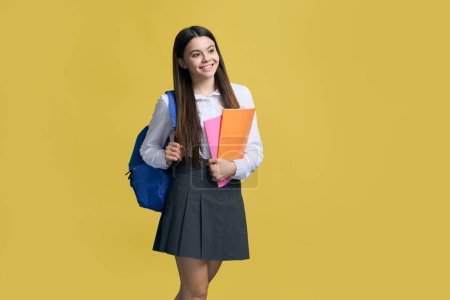 Positiv lächelndes, intelligentes Teenager-Mädchen in lässiger Schuluniform, posierend mit blauem Rucksack und Büchern, verträumt zur Seite schauend, isoliert auf gelbem Hintergrund. Zurück zum Schulkonzept. Menschen. Bildung
