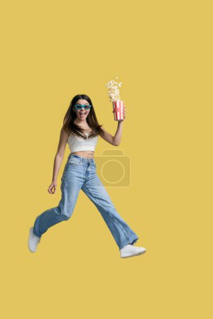 Téléchargez les photos : Adolescente excitée pleine longueur en haut blanc et en jeans en denim bleu, regardant un film sur des lunettes tridimensionnelles, exprimant des émotions positives, sautant avec une boîte pleine de pop-corn sur fond jaune - en image libre de droit