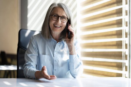 Foto de Mujer mayor sonriente confiada hablando en el teléfono móvil sentado en la oficina moderna. Concepto tecnológico - Imagen libre de derechos