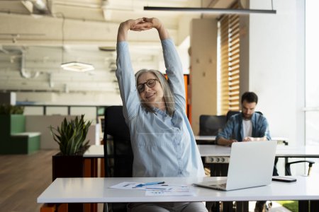Photo for Attractive smiling senior woman stretching arms and back, relaxing, taking break working in modern office. Successful confident businesswoman finish work - Royalty Free Image