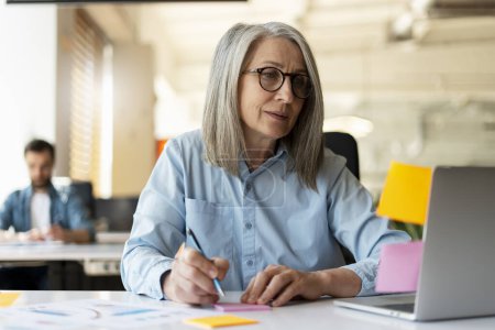 Photo for Confident senior businesswoman, writer taking notes using sticky notes looking at monitor in modern office. Gray haired manager wearing stylish eyeglasses sitting at workplace. Scrum, agile concept - Royalty Free Image