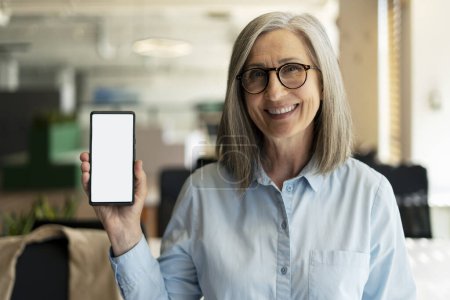 Foto de Retrato de mujer mayor sonriente atractiva sosteniendo el teléfono móvil, mostrando la pantalla digital, maqueta. Feliz 60 años de edad, mujer de pelo gris utilizando la moderna aplicación móvil de compras en línea. Concepto tecnológico - Imagen libre de derechos