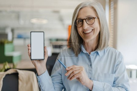 Photo for Confident smiling senior woman holding mobile phone, showing empty digital screen, mockup. Happy 60 years old, gray haired female using modern mobile app shopping online. Technology concept - Royalty Free Image
