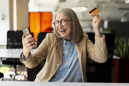 Photo for Overjoyed senior business woman holding golden credit card, mobile phone shopping online sitting in modern office. Happy entrepreneur receive payment, successful business. Mobile banking concept - Royalty Free Image