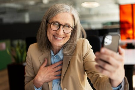 Photo for Portrait of happy gray haired senior woman holding mobile phone, communication online sitting in modern office. Video call concept - Royalty Free Image
