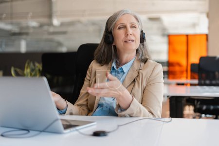 Photo for Portrait of confident gray-haired mature woman, an experienced HR manager or call center worker in formal suit and headphones, sitting at desk with laptop and looking away. People. Career. Technology - Royalty Free Image