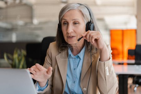 Photo for Caucasian senior gray haired stylish business woman, successful experienced manager in HR department, recruiter wearing audio headset, conducts job interviews via video link, sitting in modern office - Royalty Free Image
