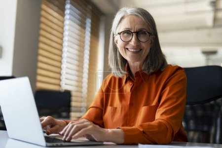 Téléchargez les photos : Portrait d'une femme d'affaires âgée souriante et confiante portant des lunettes élégantes, utilisant un ordinateur portable, travaillant en ligne dans un bureau moderne. Dactylographie financière réussie sur clavier assis sur le lieu de travail - en image libre de droit