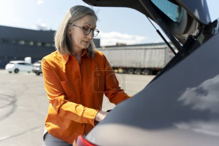 Téléchargez les photos : Portrait en gros plan d'une élégante femme âgée aux cheveux gris élégant, conductrice avec lunettes, ouvrant le coffre de la voiture, debout dans un parking à l'extérieur - en image libre de droit