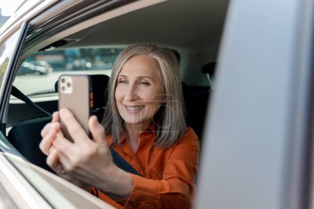 Téléchargez les photos : Souriant séduisante femme âgée tenant un téléphone mobile, prenant une photo en regardant la fenêtre assis à l'intérieur du taxi. Voyage, vacances, concept de transport - en image libre de droit
