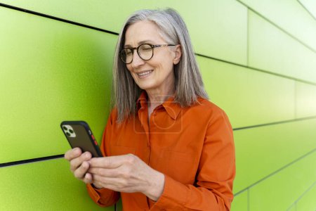 Téléchargez les photos : Portrait d'une femme mûre aux cheveux gris souriant tenant un smartphone à l'aide d'une application mobile faisant du shopping en ligne debout dans la rue. Joyeux 60 ans femme lisant le message texte, communication. Technologie - en image libre de droit