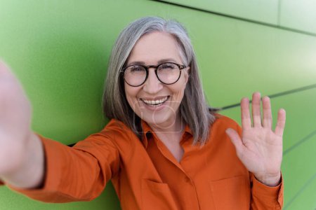 Happy grey haired senior woman taking selfie, waving hand, communication online, having video call standing outdoor. Smiling influencer recording video on the street. Technology concept 